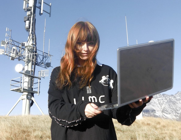 Ninajirachi standing in a field with a laptop, looking at the camera
