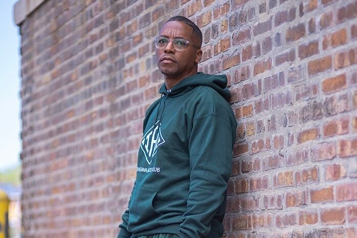 Lupe Fiasco wearing a green outfit, leaning against a brick wall