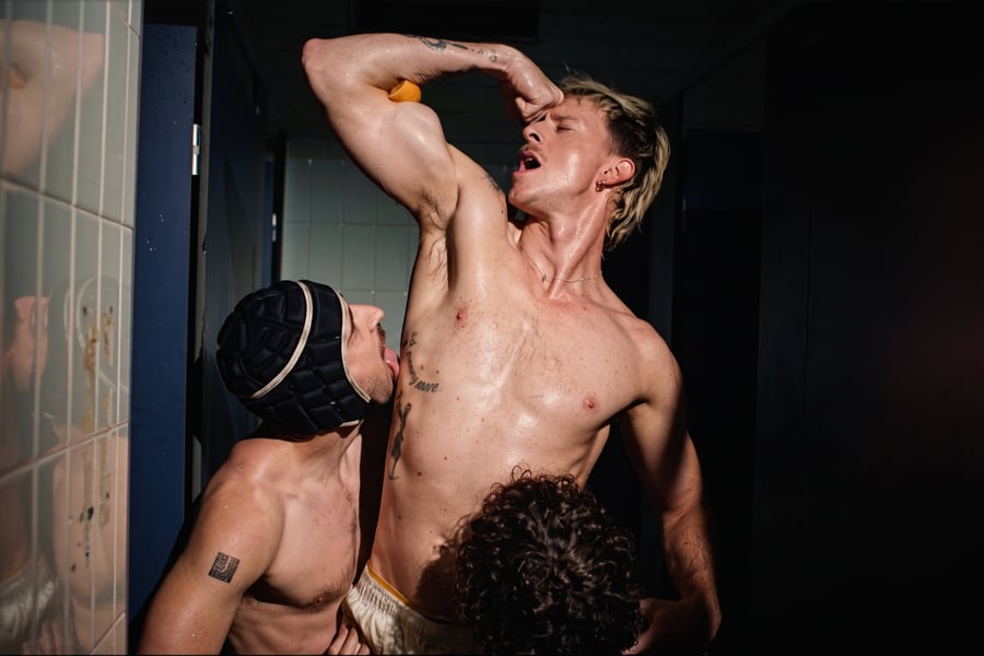A man raising his arm beside two other men in a tiled shower room.
